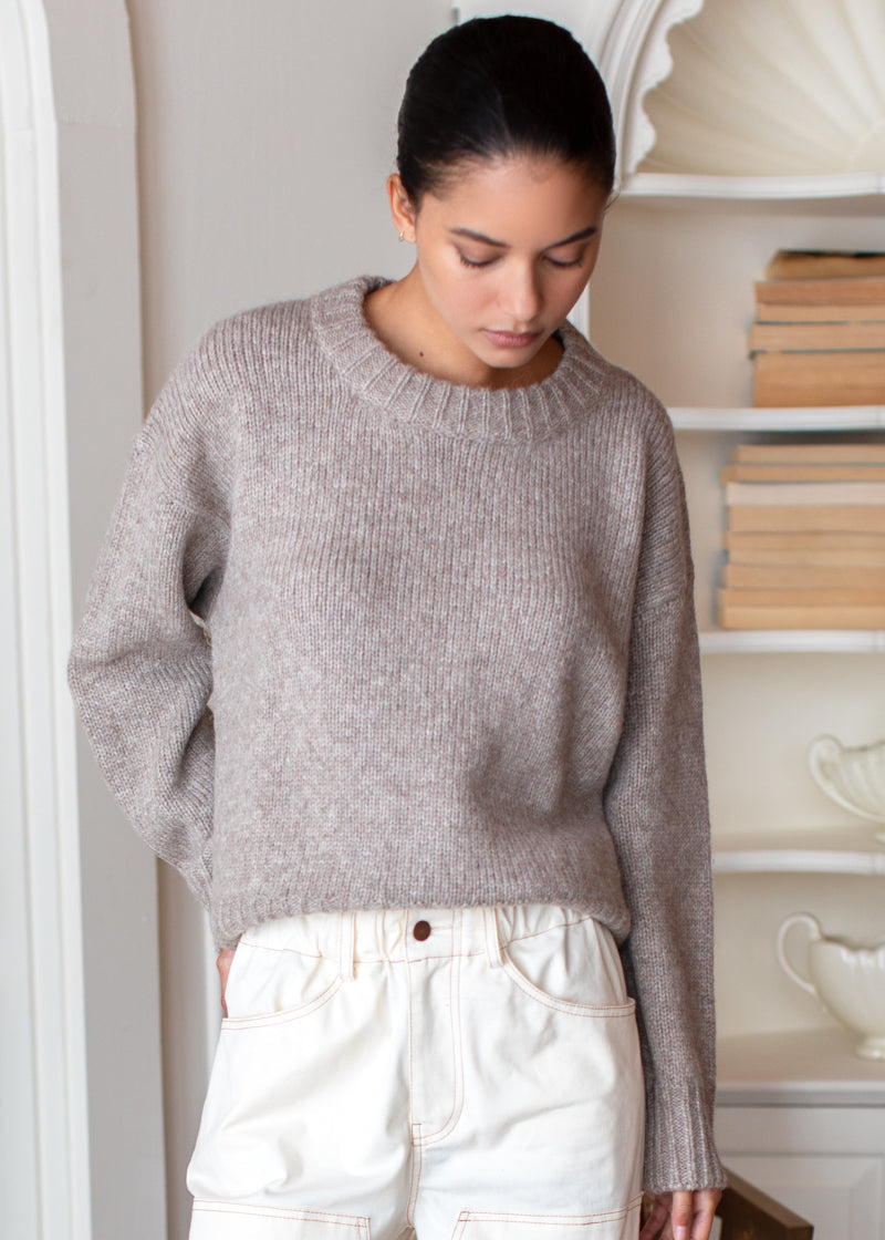 A woman wearing a light taupe ribbed knit crewneck sweater and off-white high-waisted jeans, standing in a bright, neutral-toned room with bookshelves in the background.