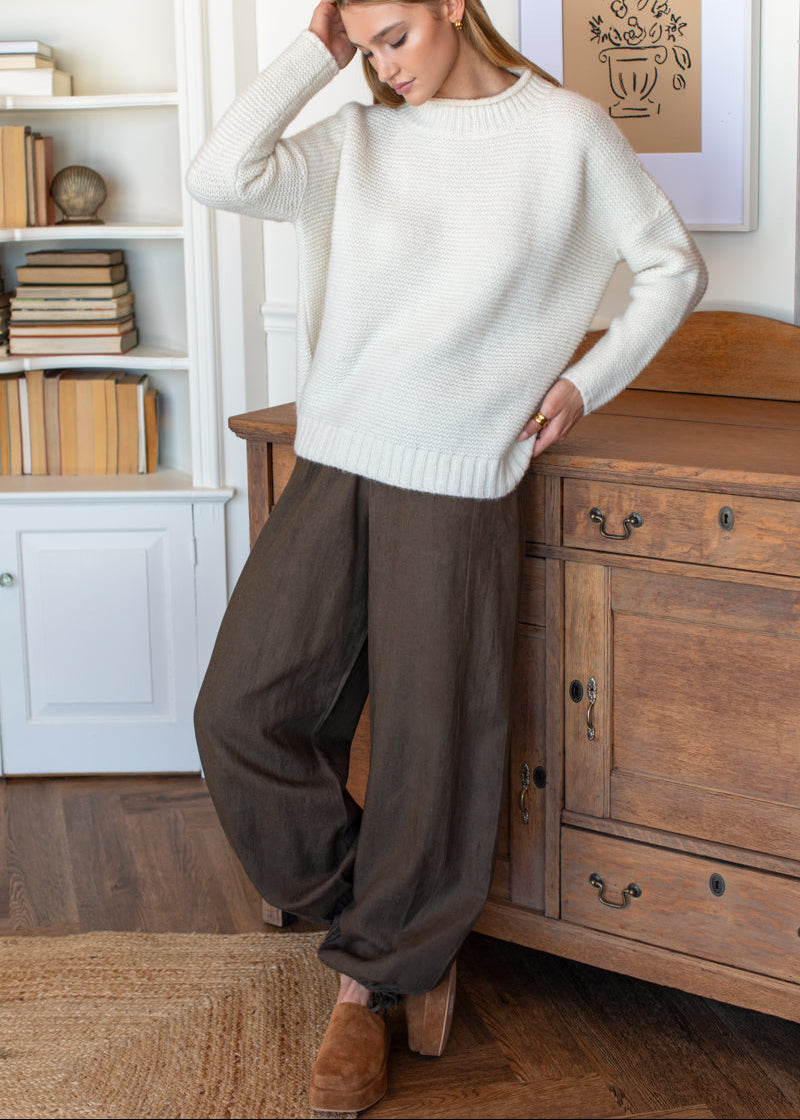 A woman stands in a cozy room wearing a cream knit crewneck sweater and dark brown wide-leg pants, leaning against a wooden cabinet.