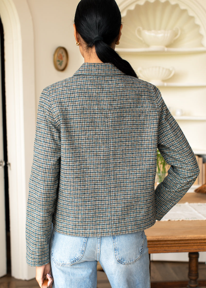 A person shown from behind wearing a cropped, long-sleeve houndstooth blazer in muted gray, beige, and brown tones, paired with light blue jeans in an indoor room setting.