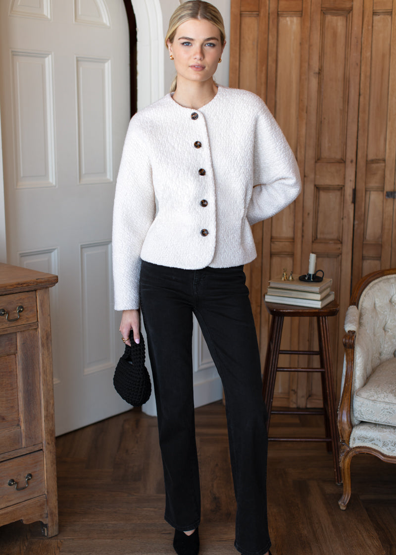 Woman wearing a white jacket and black pants in a room with wooden furniture.