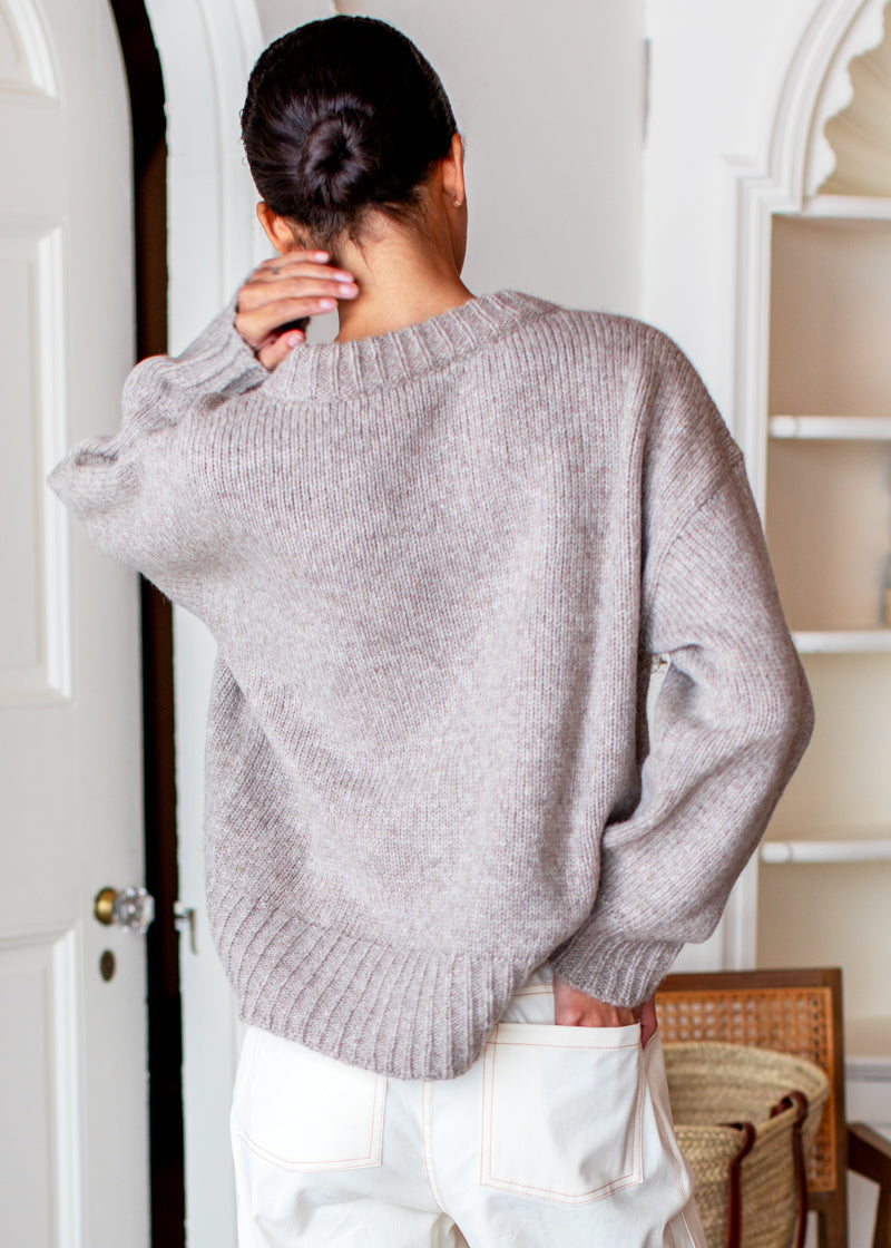A person shown from behind wearing a light taupe knit sweater and white pants in an indoor room setting.
