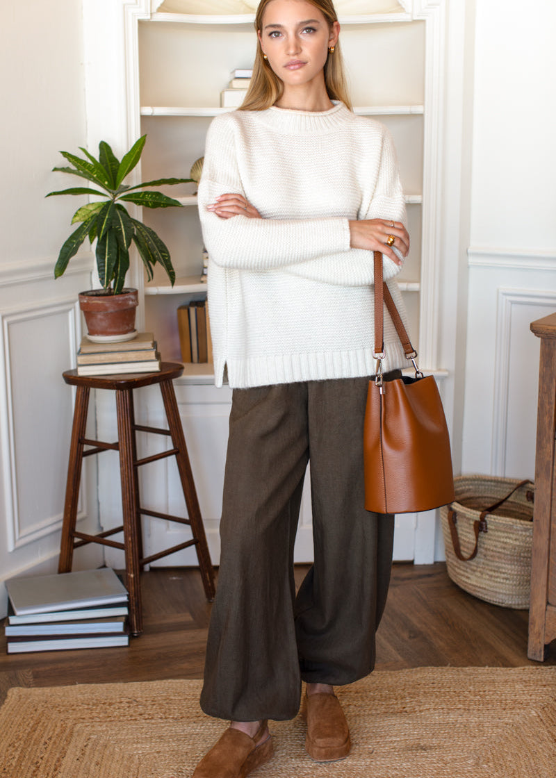 A woman stands in a bright indoor room wearing a cream knit sweater, wide-leg dark brown trousers, and brown suede shoes, while holding a brown leather bucket bag.