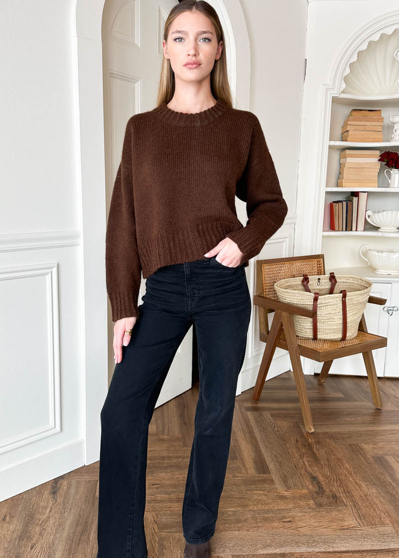 A woman modeling a brown crewneck knit sweater and dark blue jeans in a bright, white-walled room with a wooden floor and a woven basket bag on a chair.