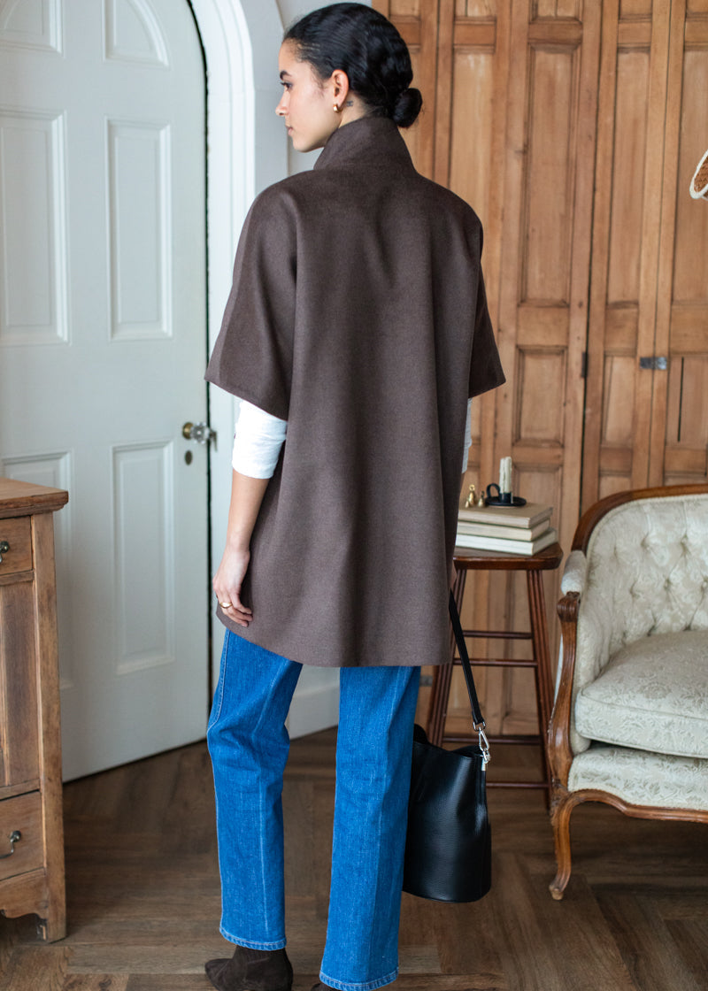 A woman stands with her back to the camera wearing a brown sleeveless wrap or poncho over a white long-sleeve top, blue jeans, and brown ankle boots, holding a black shoulder bag in a rustic indoor room with wood paneling and furniture.