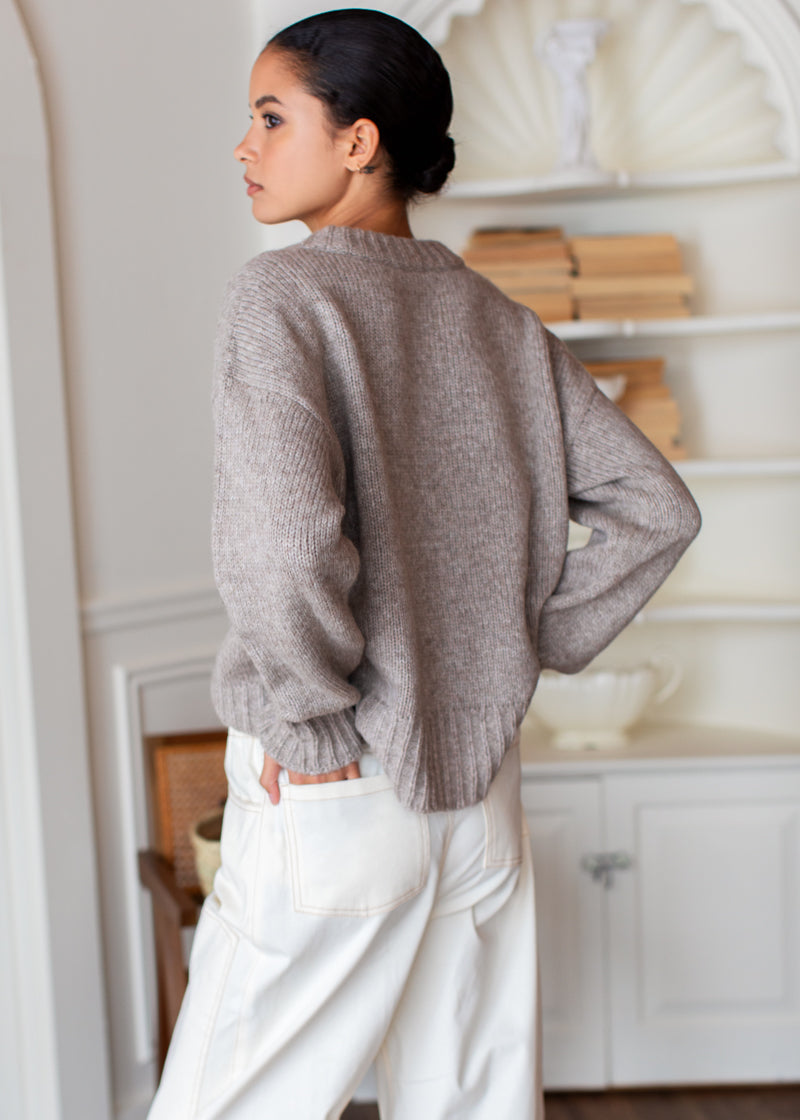 A woman wearing a taupe knit sweater and white wide-leg pants, photographed from behind in a bright, white interior room.