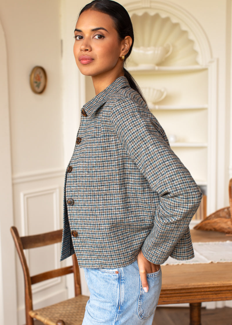 A woman wearing a cropped, long-sleeve plaid button-up jacket in blue, beige, and brown tones, paired with light-wash blue jeans, standing in a warm, classic indoor room with a wooden chair and table in the background.