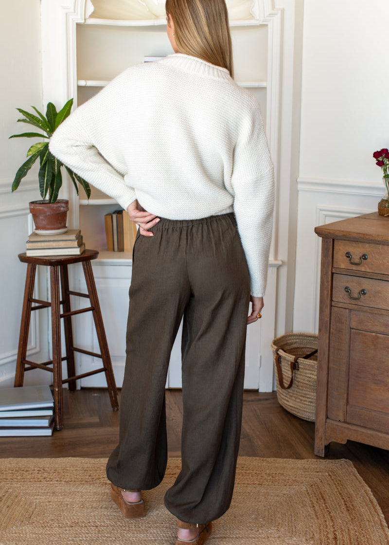 A woman shown from the back wearing a cream knit sweater and dark brown wide-leg trousers in a cozy, neutral-toned room with wooden furniture and a bookshelf.