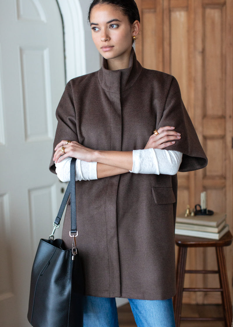 A woman wearing a brown cape-style coat over a white long-sleeve top and blue jeans, holding a black leather bucket bag. She is standing in an indoor setting with a white door on the left and a wooden cabinet in the background.
