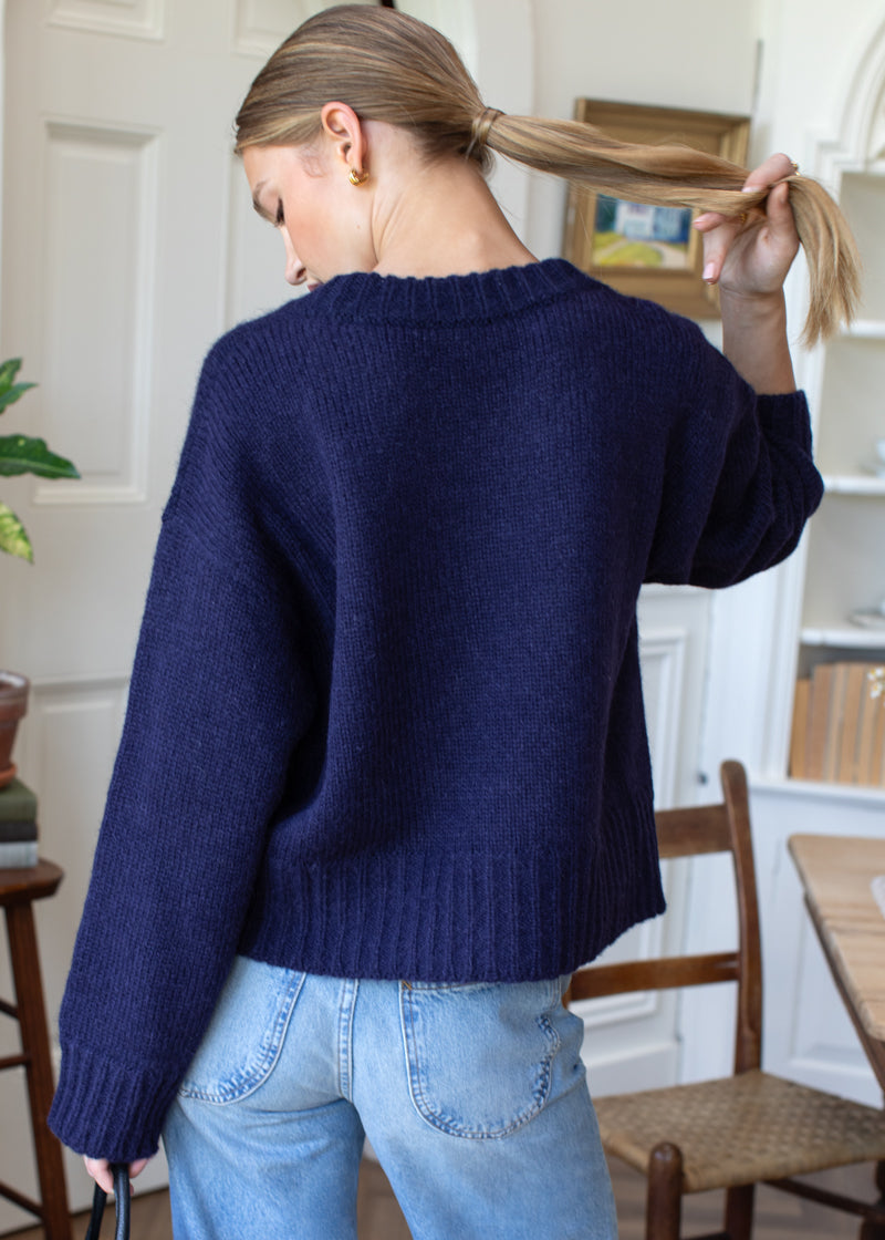 A woman viewed from behind wearing a navy blue ribbed knit sweater and light blue jeans in a bright indoor room.