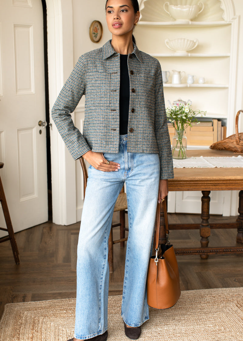A woman stands indoors wearing a gray and black houndstooth button-up jacket over a black top, light-wash wide-leg jeans, and dark flats. She holds a tan leather handbag beside a wooden table in a styled room with shelves and a rug.