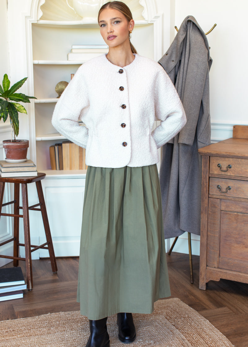 Woman wearing a white jacket and green skirt standing in a room with wooden furniture and decor.