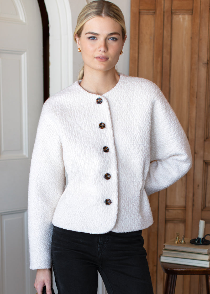 Woman wearing a white textured jacket with brown buttons indoors.