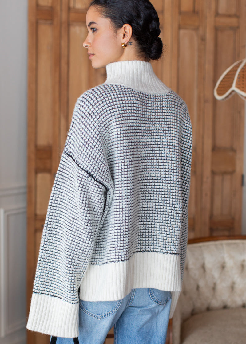 A woman shown from the back wearing an oversized knit sweater with a black and white micro-check pattern, a solid white ribbed turtleneck, and white ribbed cuffs and hem, paired with light blue jeans in an indoor setting.