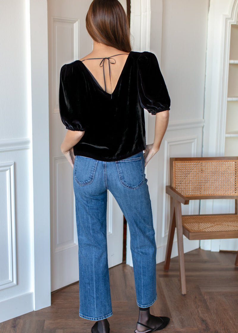 Person wearing a black top and blue jeans standing in a room with wooden flooring and white walls.