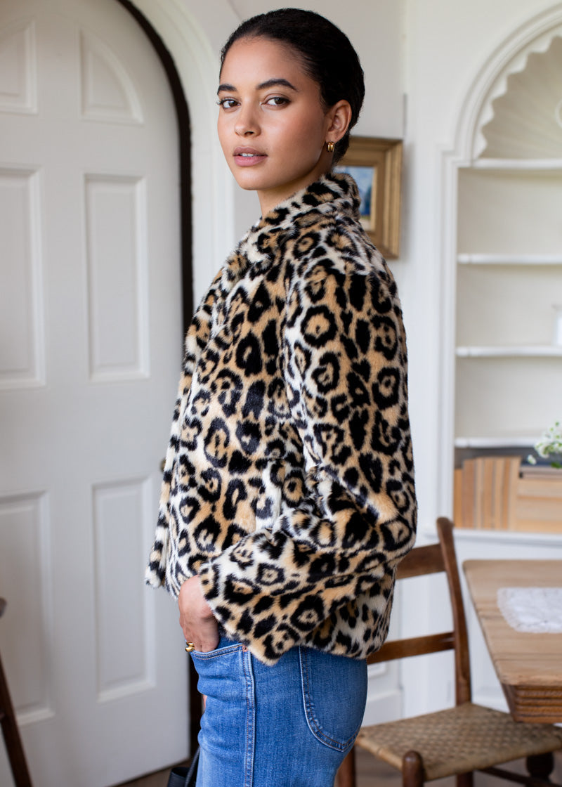 A woman wearing a plush leopard-print faux fur jacket and blue jeans, standing in a bright, neutral-toned indoor room.