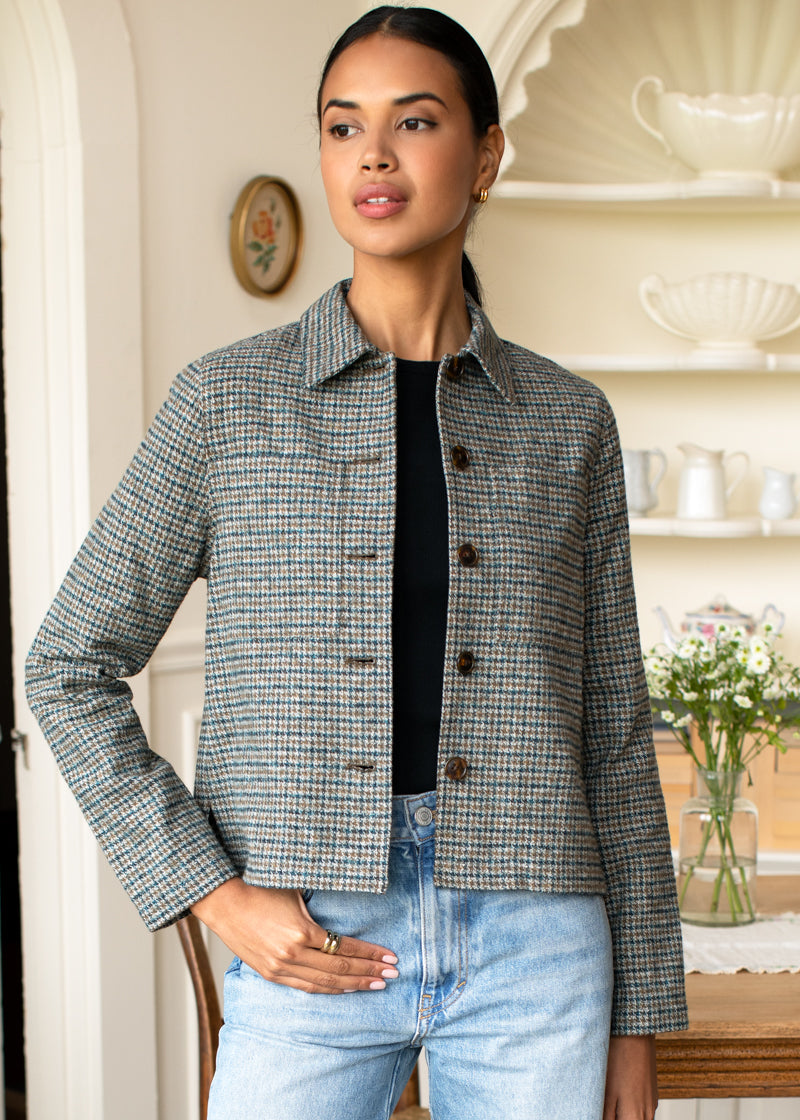 A woman modeling a cropped, long-sleeve houndstooth blazer in gray and black, worn over a black top and light blue jeans in a warm, cream-colored indoor setting.