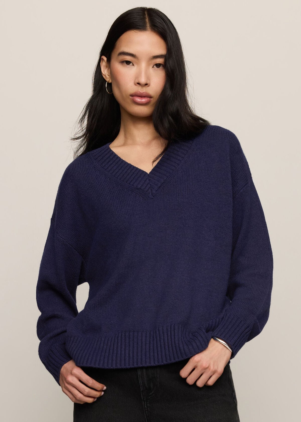 A woman wearing a navy blue knit V-neck sweater with dropped shoulders and ribbed cuffs, paired with black jeans, against a plain light beige studio background.