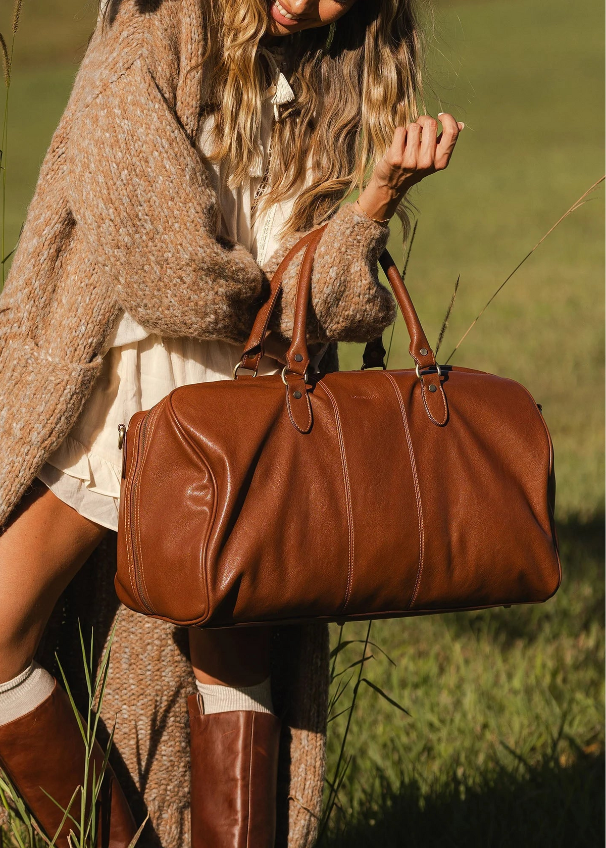 Person holding a brown leather duffel bag in a grassy field