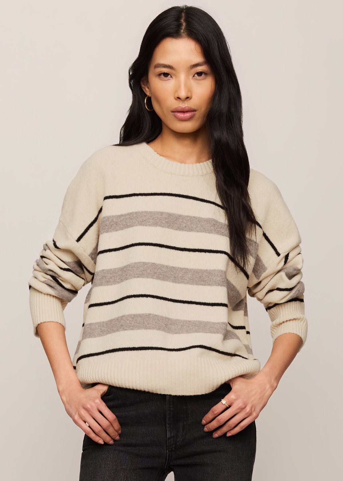 A woman modeling a cream-colored knit sweater with horizontal black and gray stripes, paired with black jeans, against a plain light beige studio background.