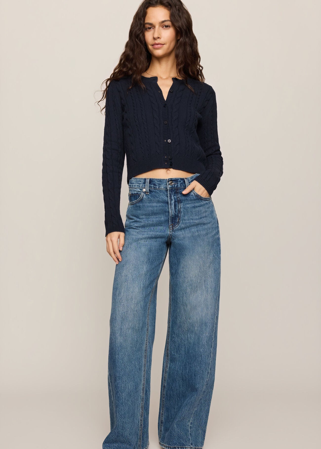 A woman modeling a dark navy blue cropped button-up knit top and high-waisted, wide-leg blue denim jeans against a solid light beige studio background.